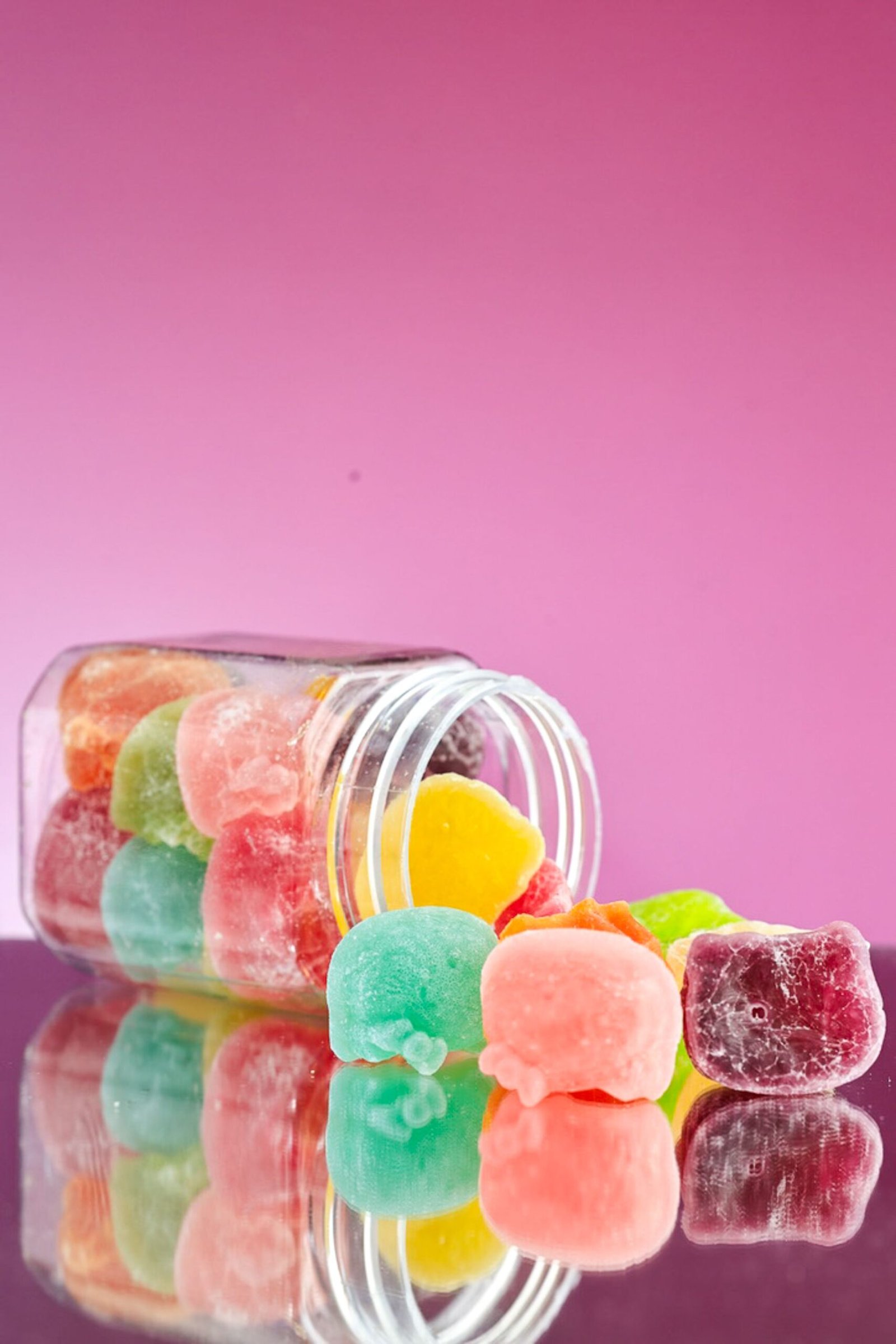 A glass jar of assorted gummy candies spills onto a reflective surface with a pink background.